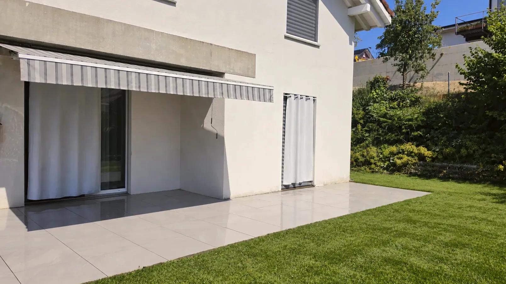 Terrasse en grès cérame devant une maison blanche à store banne, bordée d'une pelouse fraîchement tondue